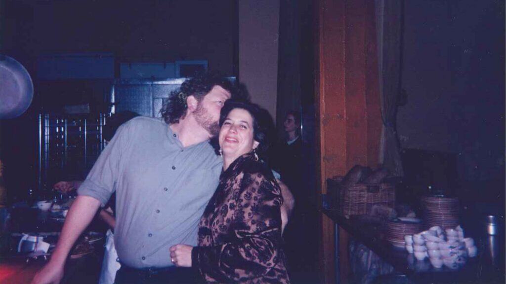 A man kisses a woman on the cheek as they stand close together in a dimly lit room with kitchenware and baskets in the background.