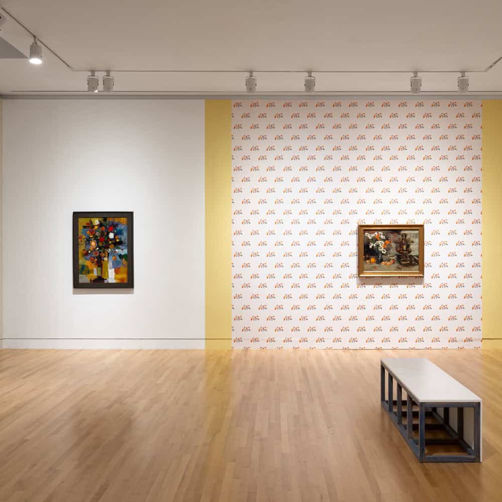 A gallery room with two framed paintings of flowers on adjacent walls, a bench on the wooden floor, and patterned wallpaper behind one artwork.