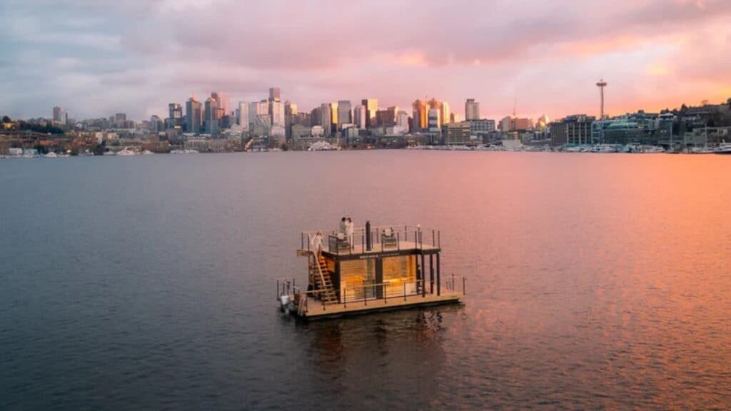 A small floating house sits on a calm body of water, with a city skyline and the Space Needle in the background at sunset.