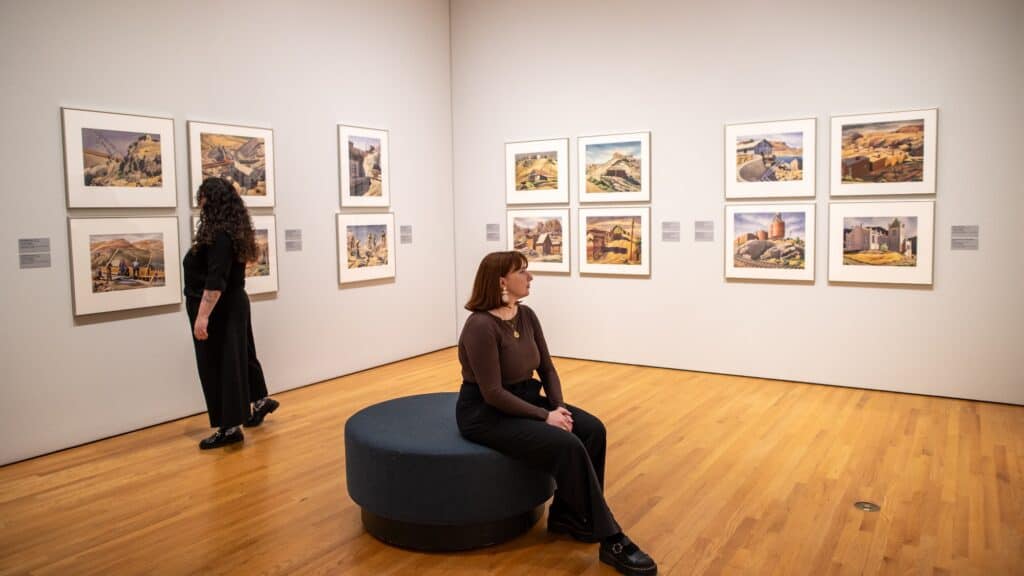 Two women in a gallery room view paintings on white walls; one sits on a round bench while the other examines artwork closely.