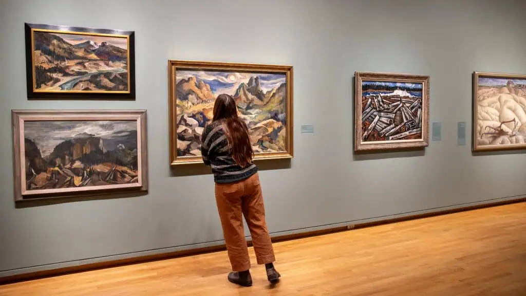 A person stands closely observing a landscape painting in an art gallery, surrounded by several framed paintings on a light gray wall.