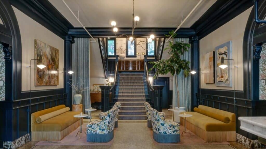 Chic hotel lobby with mustard sofas, floral chairs, round tables, contemporary art on walls, and a staircase leading to an upper floor. Modern lighting hangs from the ceiling.