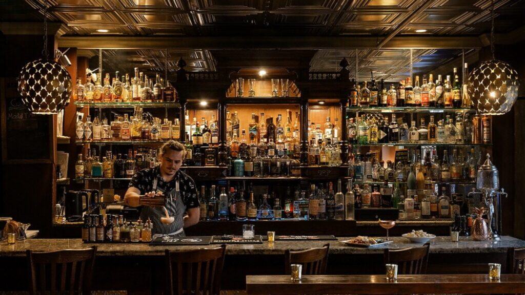 Bartender prepares a drink behind a dimly lit bar with shelves full of various liquor bottles; barstools and drinks are visible in the foreground.