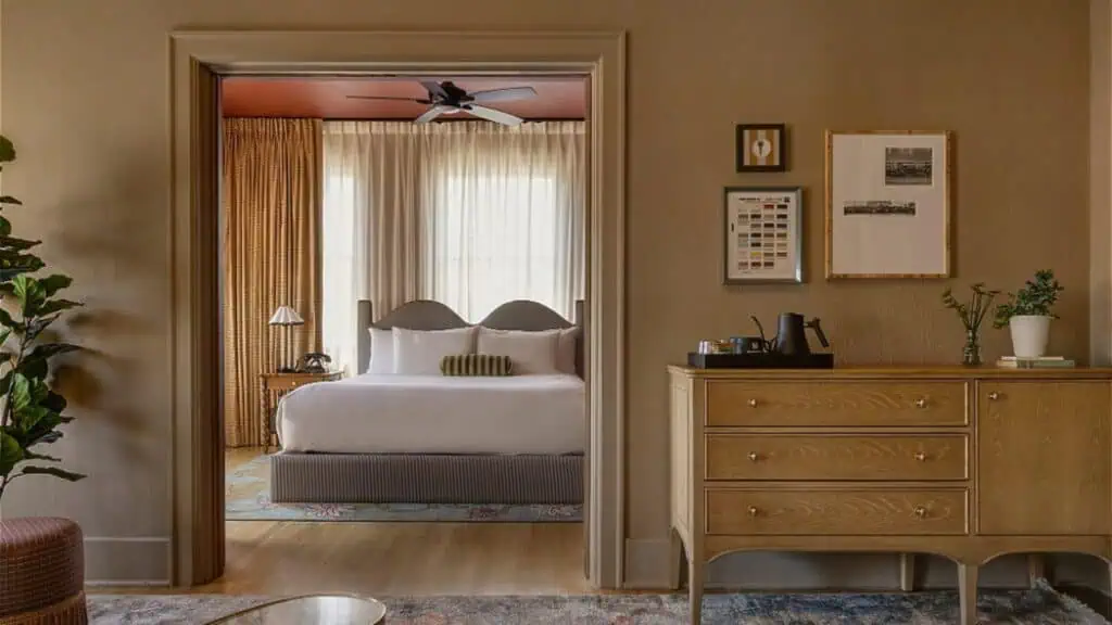 A tidy bedroom with a double bed, large windows with sheer curtains, and a ceiling fan, viewed from an adjacent room with a wooden dresser and framed wall art.