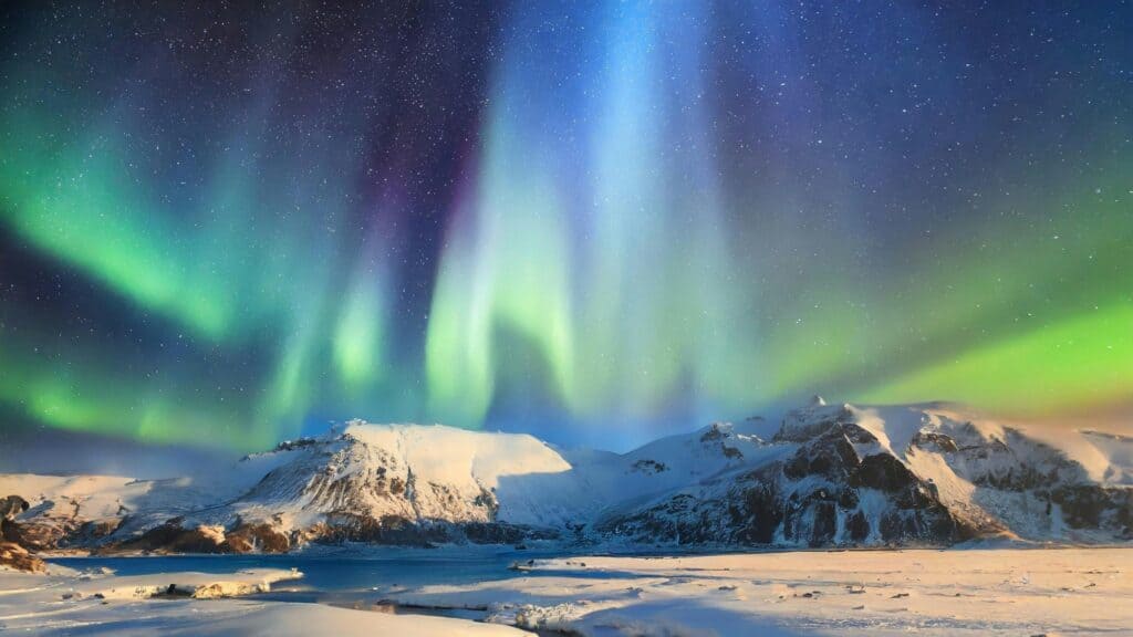 Northern lights in green and blue hues illuminate a night sky above snow-covered mountains and a frozen landscape.