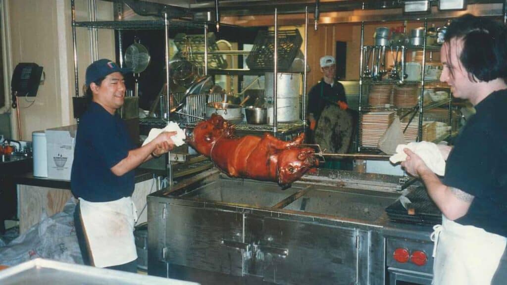 Two chefs in a commercial kitchen are handling a whole roasted pig on a spit over a metal roasting pit, while another person works in the background.
