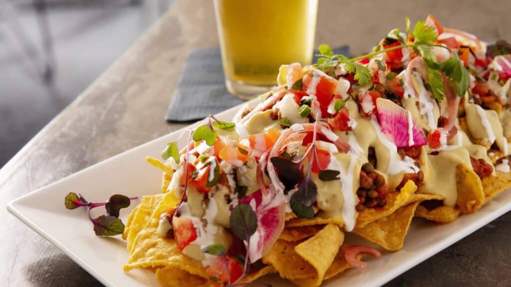 A plate of nachos topped with ground meat, diced tomatoes, onions, creamy sauces, and fresh herbs—a perfect Seattle game day food—sits in front of a glass of beer in the background.
