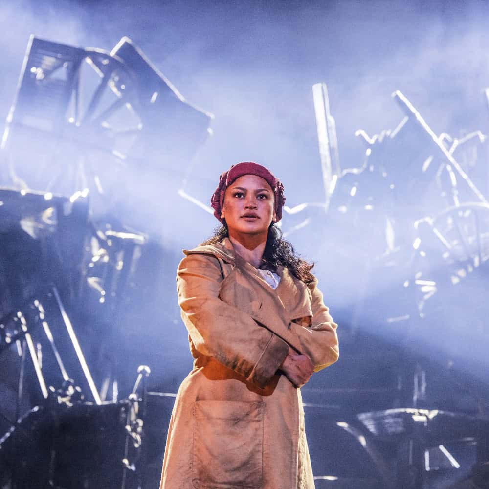 A woman in a tan coat and red headscarf stands with arms crossed on a dimly lit stage, with stacked chairs and dramatic lighting in the background.
