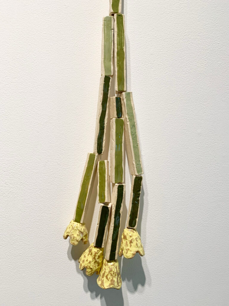 A ceramic wall sculpture by Gabriel Stromberg, resembling a bundle of green vegetable stalks with yellow, bulb-like ends, hangs vertically against a white wall—a striking piece from the Studio Sessions collection.