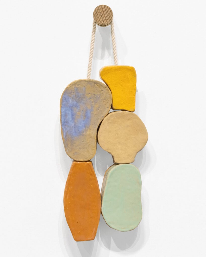 Five irregularly shaped, colorful ceramic pieces are stacked vertically and hung from a rope on a wall-mounted hook.