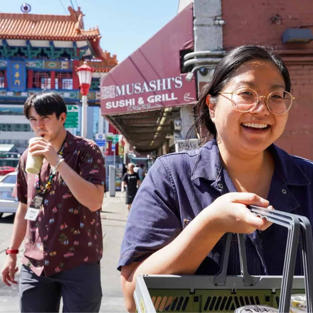 Two people walk outdoors near Musashi’s Sushi & Grill in Seattle; one smiles while carrying a basket, the other drinks from a cup. A colorful building in the background adds to the vibrant scene as they celebrate AANHPI Month.