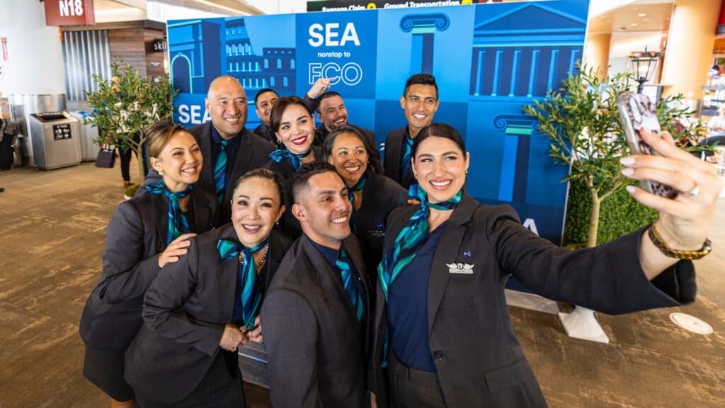 A group of airline crew members in uniform poses for a selfie in front of a sign indicating flights from Seattle to Rome inside an airport terminal.