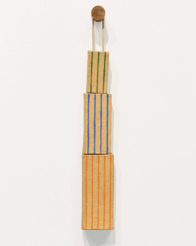 Three vertically stacked, textured rectangular blocks with colored vertical stripes (green, blue, orange) hang from a cord attached to a round, woven element on a plain white wall.