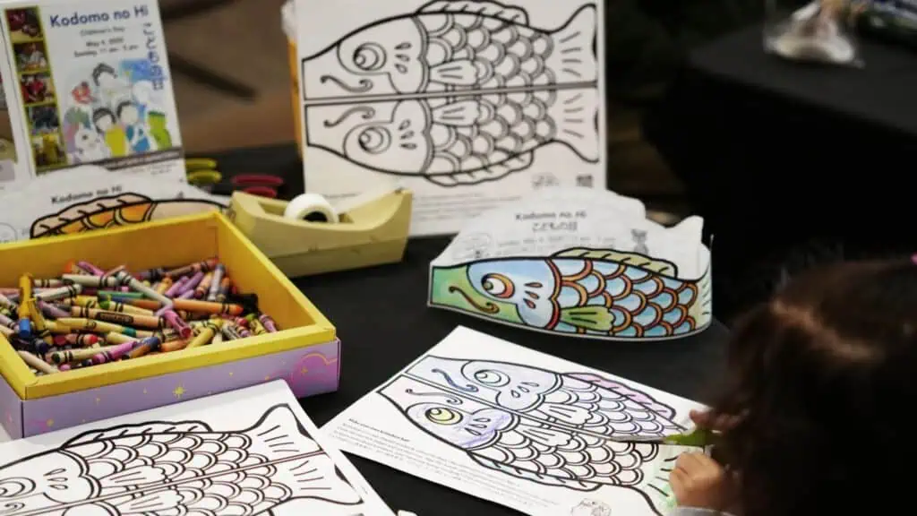 A child colors a fish-shaped paper at a table with crayons, alongside completed fish coloring pages and Kodomo no Hi flyers—capturing the festive spirit often seen at Spring Festivals Washington.