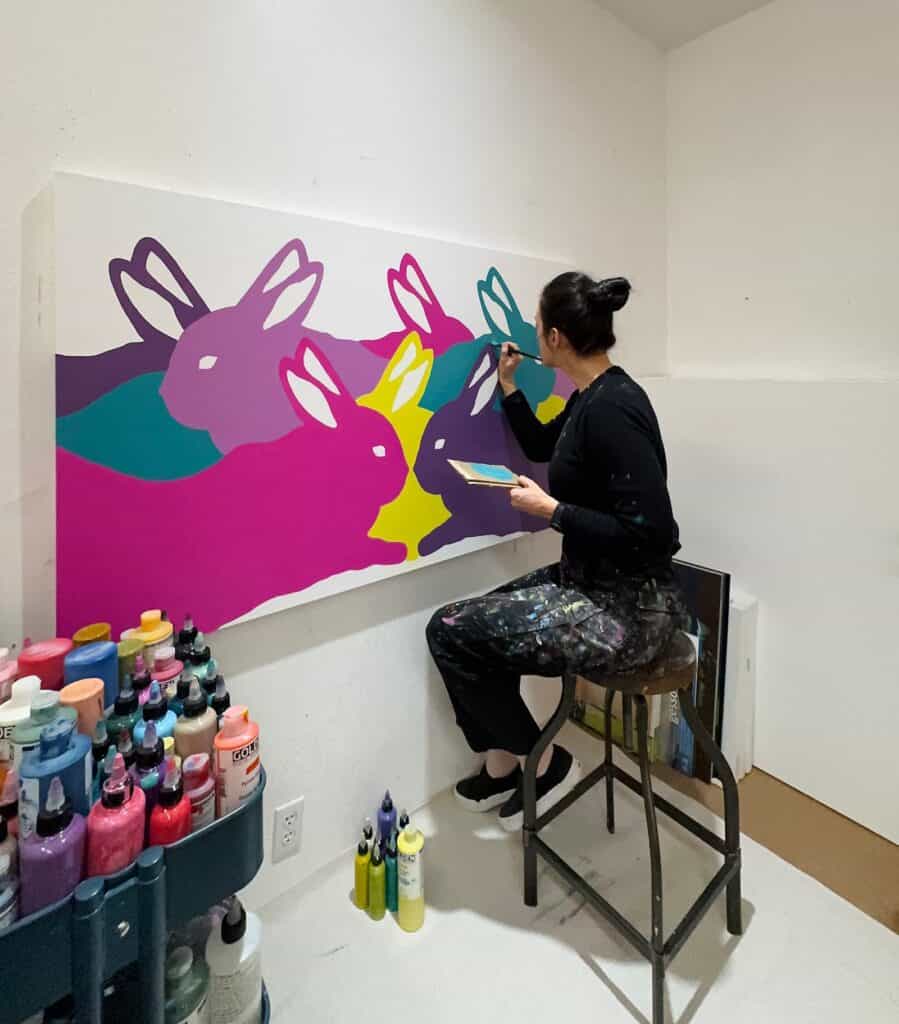 A woman sits on a stool painting colorful overlapping bunny silhouettes on a large canvas in a studio with paint bottles nearby.