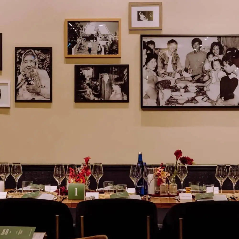 A dining table set with glassware and red floral centerpieces sits beneath a wall decorated with framed black and white photographs.