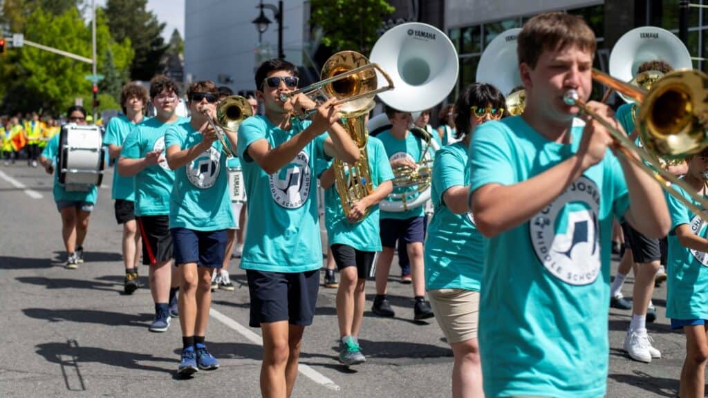 A high school marching band in teal shirts plays brass and percussion instruments while walking down a street during one of the vibrant Washington Spring Festivals.