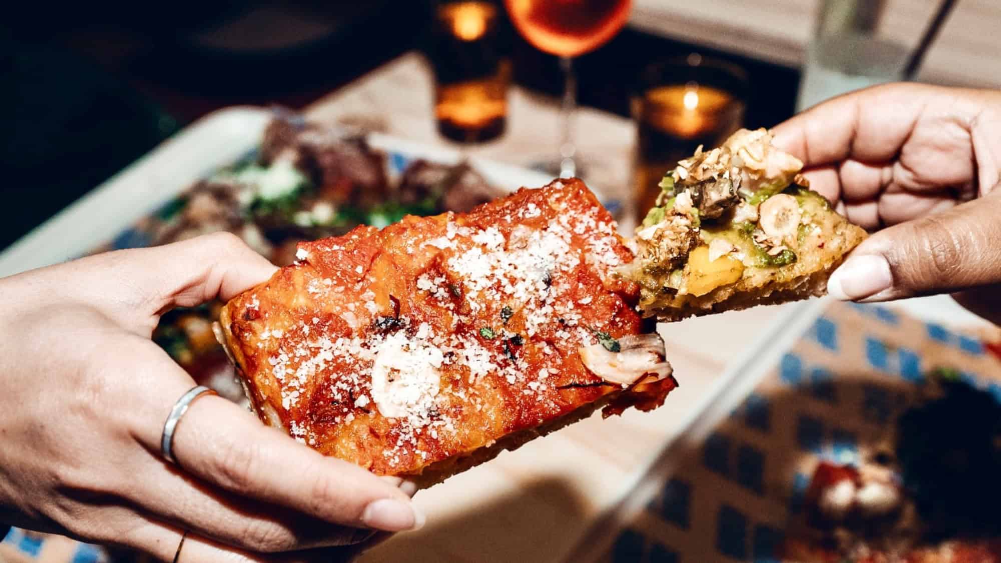 Two hands holding different slices of pizza, one with tomato sauce and cheese, the other with assorted toppings, over a table with drinks and food.