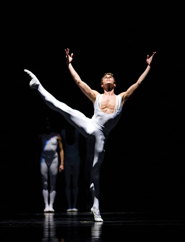 A male ballet dancer in a white outfit performs a high leg extension on stage with Pacific Northwest Ballet, another dancer blurred in the background against a dark backdrop.