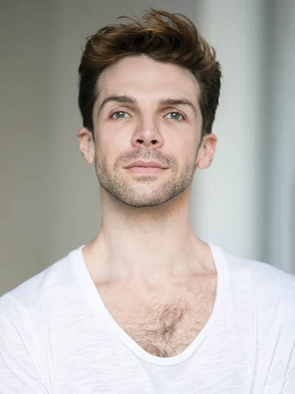 A young man with light skin, short brown hair, and blue eyes wears a white shirt and looks directly at the camera with a neutral expression, reflecting the poise often seen in Pacific Northwest Ballet portraits.