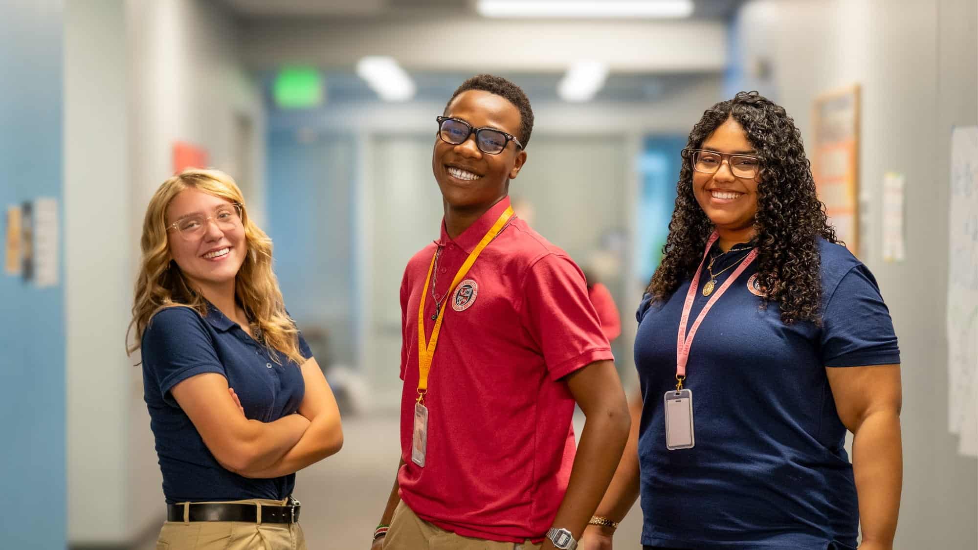 Three students wearing uniforms and ID badges stand smiling in a brightly lit hallway with blue walls, reflecting the positive early outcomes seen at Washington charter public schools.