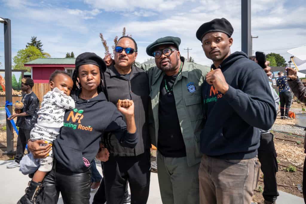 Four adults, one holding a child, stand together outdoors, facing the camera with raised fists. They are wearing casual and military-style clothing. It appears to be a group event.