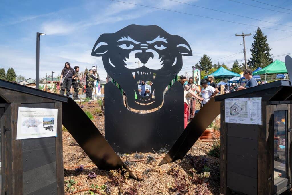 Large black metal sculpture of an open-mouthed animal head at an outdoor event, with people and green tents in the background.