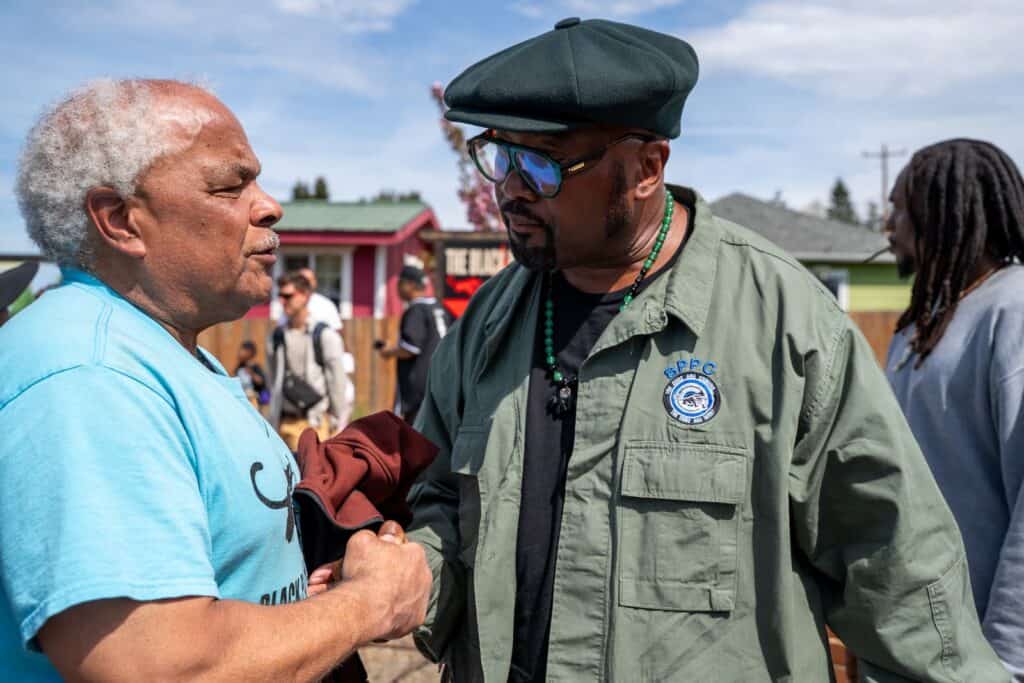 Two men stand outdoors, shaking hands and talking at an event; one wears a blue shirt, the other a green jacket and beret. Other people and houses appear in the background.