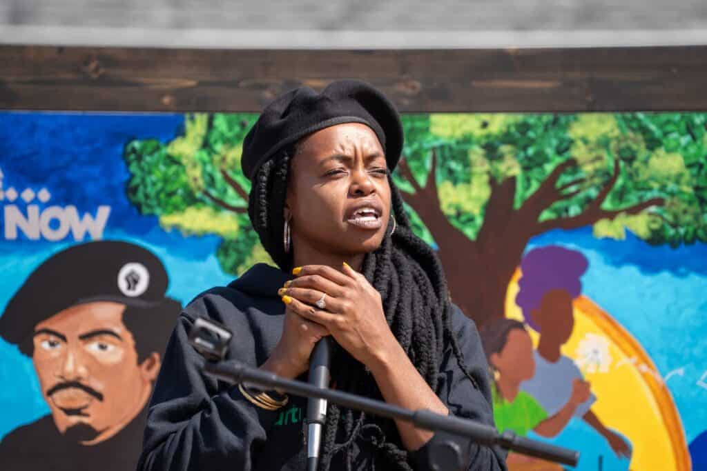 A person with long braids and a black beret speaks into a microphone at Black Panther Park in the World, standing before a colorful mural depicting people and a tree.