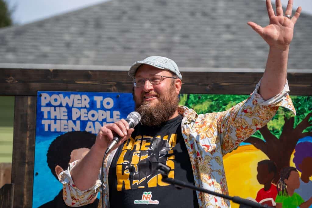 A bearded man in glasses and a cap speaks into a microphone and waves, standing in front of a colorful mural and sign reading "POWER TO THE PEOPLE THEN AND.
