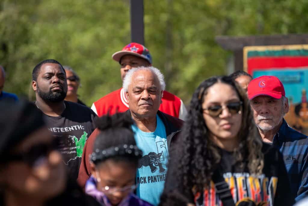 A diverse group of people stands outdoors, some wearing sunglasses and hats, with greenery and a colorful sign visible in the background.