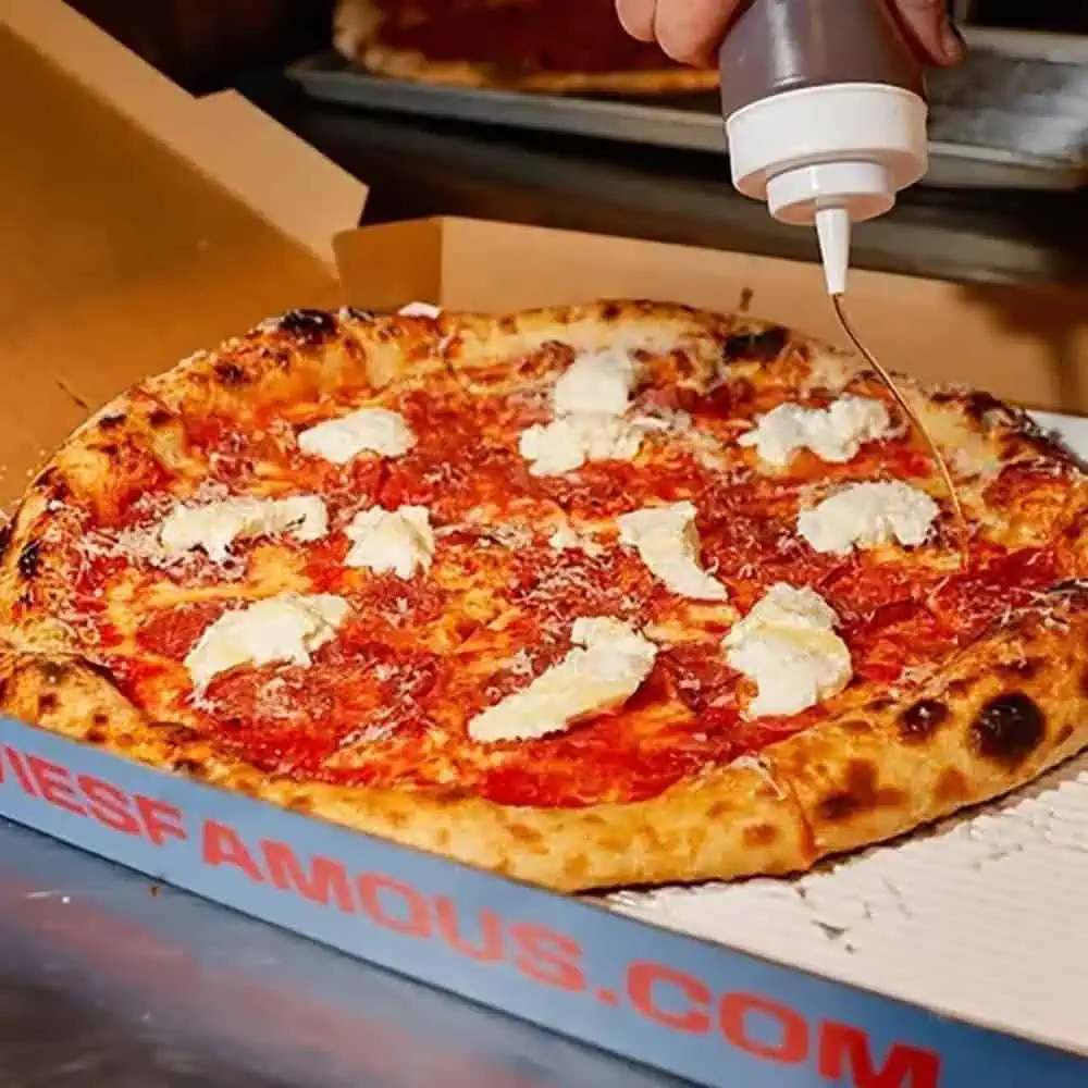 A hand drizzles sauce from a squeeze bottle onto a pizza topped with cheese and tomato sauce, in an open cardboard box—a mouthwatering take on Seattle pizza trends.