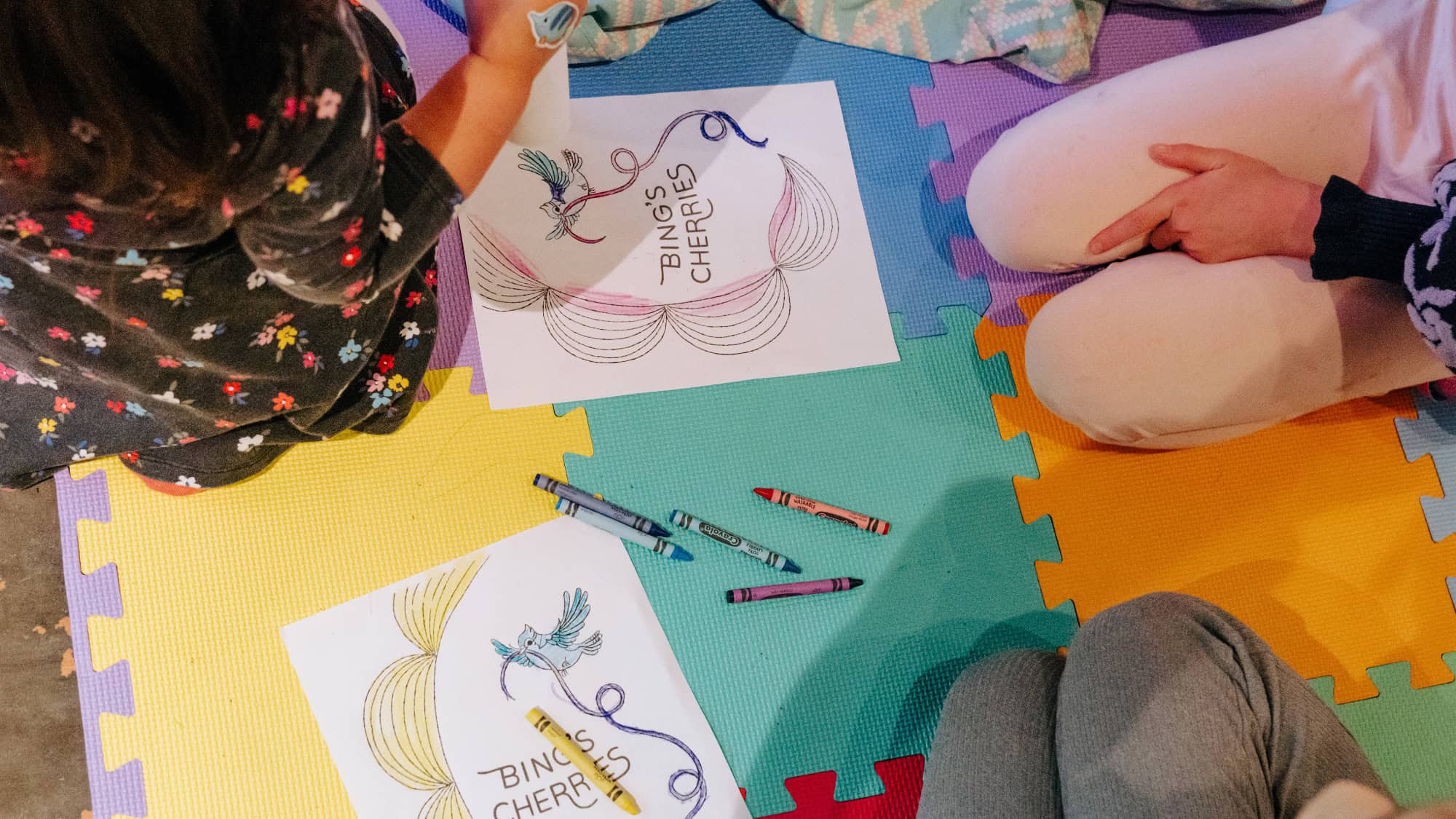 Two children sit on colorful foam mats, coloring "Bingo's Cherries" activity sheets with crayons. Their faces are not visible.