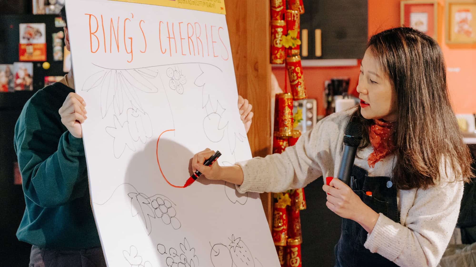 A woman holding a microphone draws on a large paper labeled "Bing's Cherries" while another person holds the paper.