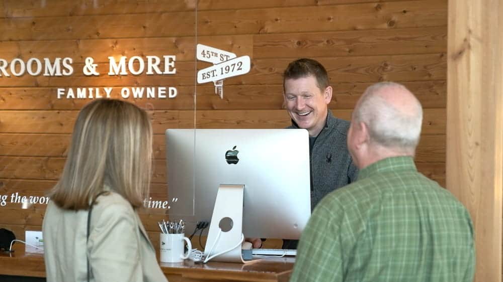 A man stands behind a counter with an iMac, smiling and talking to a woman and an older man at Bedroom and More, a family-owned business known for its mattresses with natural materials.
