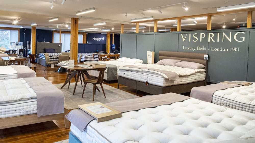 Showroom with multiple luxury beds on display, wooden floors, and a Vispring sign on the wall; tables and chairs are placed among the beds.
