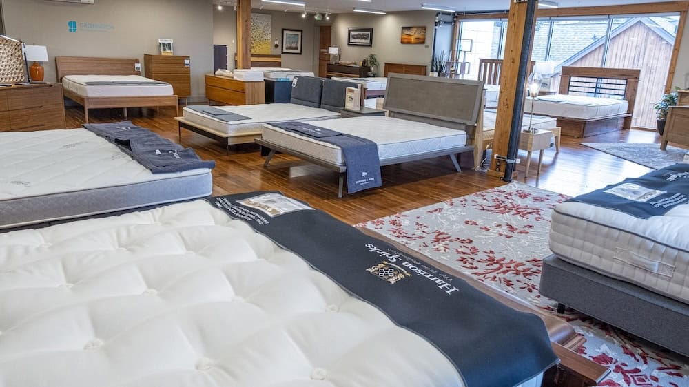 A showroom with multiple beds and mattresses on display, wooden flooring, modern decor, and natural light coming through large windows.