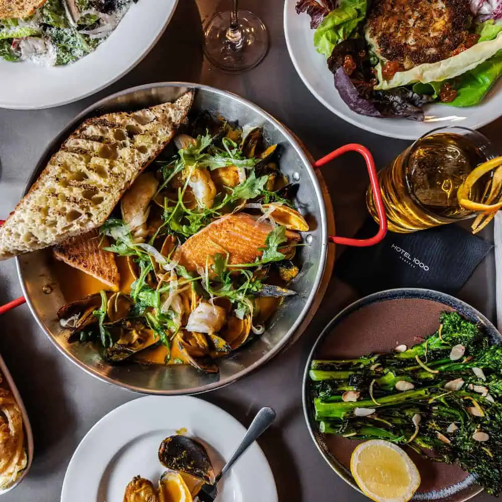 A table with assorted dishes including grilled fish with mussels and bread, broccolini, a patty with salad, a glass of drink with lemon, and a napkin labeled "Hotel Rodol".