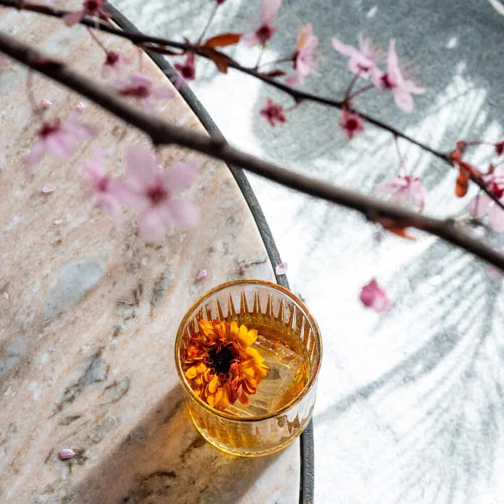 A glass of amber-colored liquid with a yellow flower is placed on a marble surface, with a branch of pink blossoms visible in the foreground.