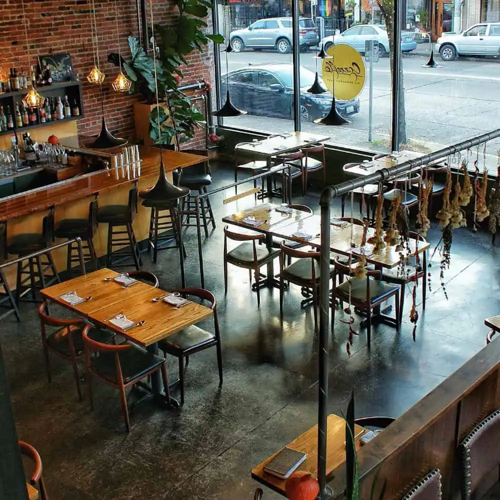 A modern restaurant interior with wooden tables, chairs, a bar area, hanging lights, large windows, and dried herbs suspended from the ceiling.
