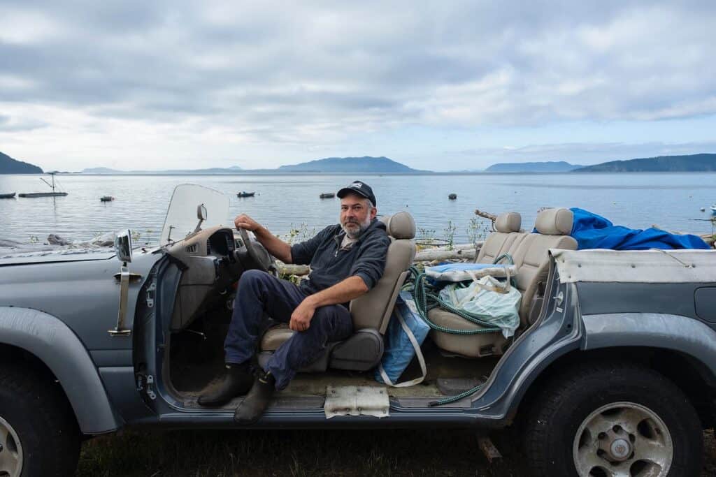 A man sits in a convertible filled with various items, parked by calm Legoe Bay, with mountains and boats—possibly Reefnetters—in the background beneath a cloudy sky, capturing a moment fit for a photo essay.