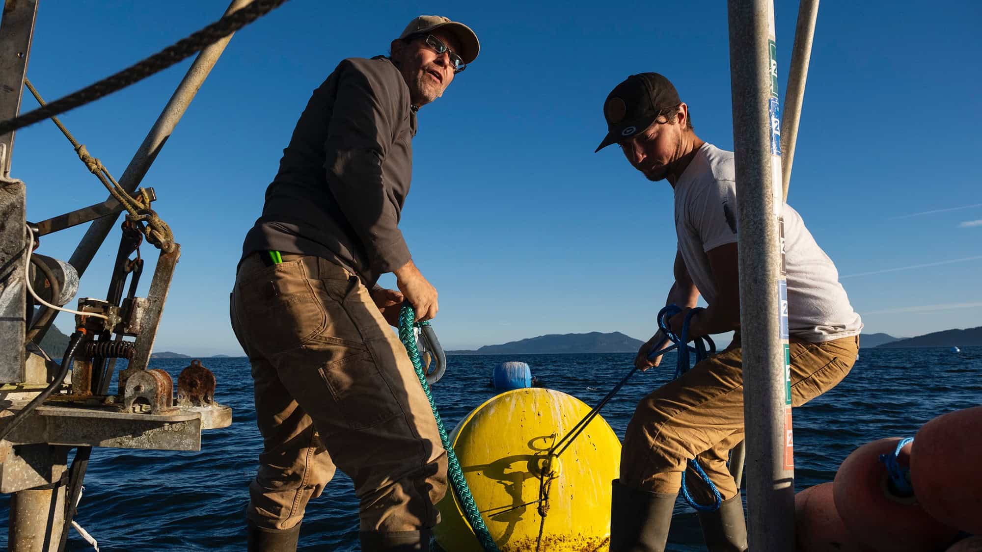 Two people on a boat handle ropes attached to a large yellow buoy, with the ocean and distant mountains in the background under a clear sky.