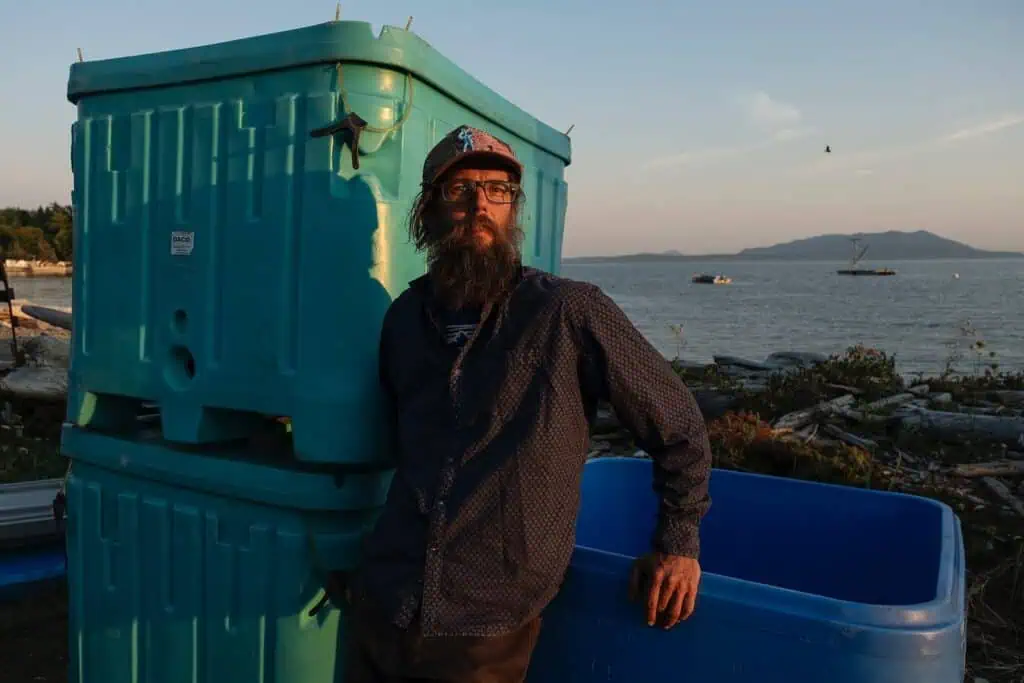 A bearded man wearing glasses and a cap leans against large blue bins near a shoreline, with water, boats, and mountains visible in the background.
