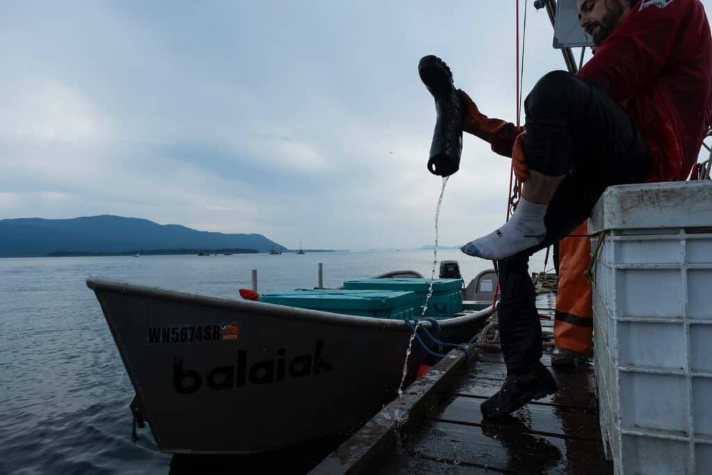 A person sitting on a dock pours water out of a black boot, with a boat labeled "balaiak" and mountains visible in the background.