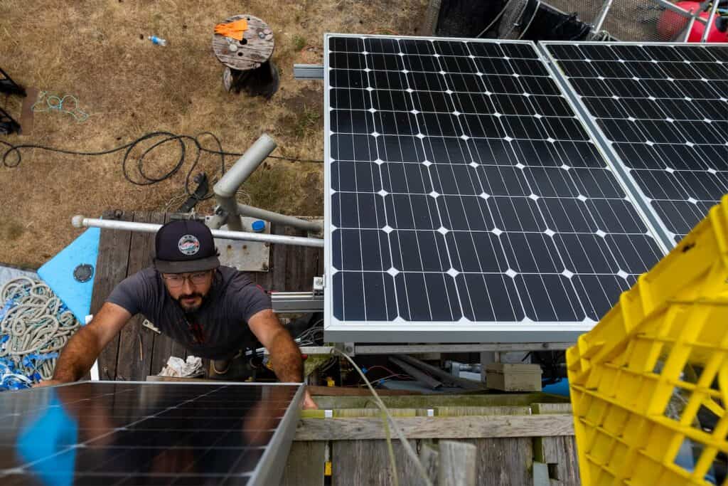 A man adjusts solar panels on a rooftop, with tools and equipment scattered around him. The scene is viewed from above.