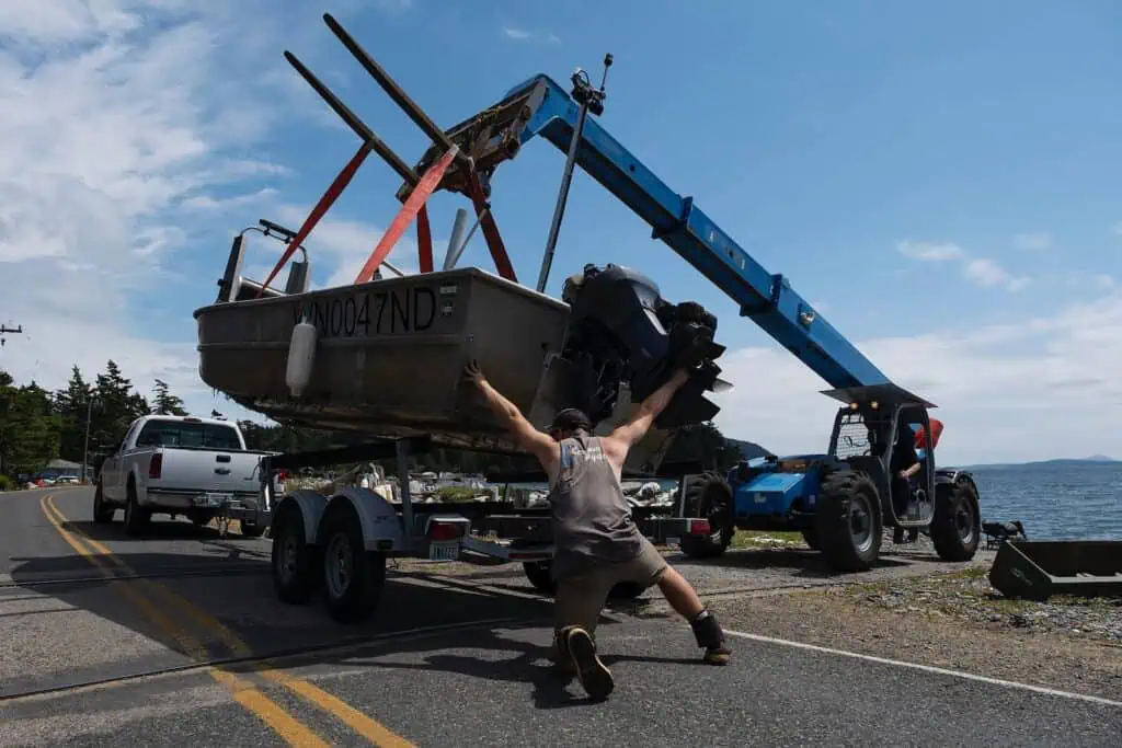 A man stabilizes a small boat on a trailer while a blue forklift lifts it beside a white pickup truck on a coastal road.