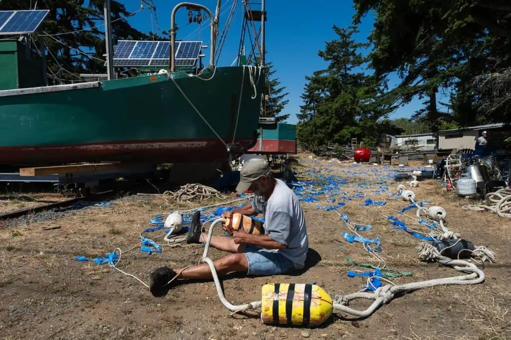A man sits on the ground assembling buoys and ropes near a green boat surrounded by scattered fishing equipment and trees in an outdoor area.