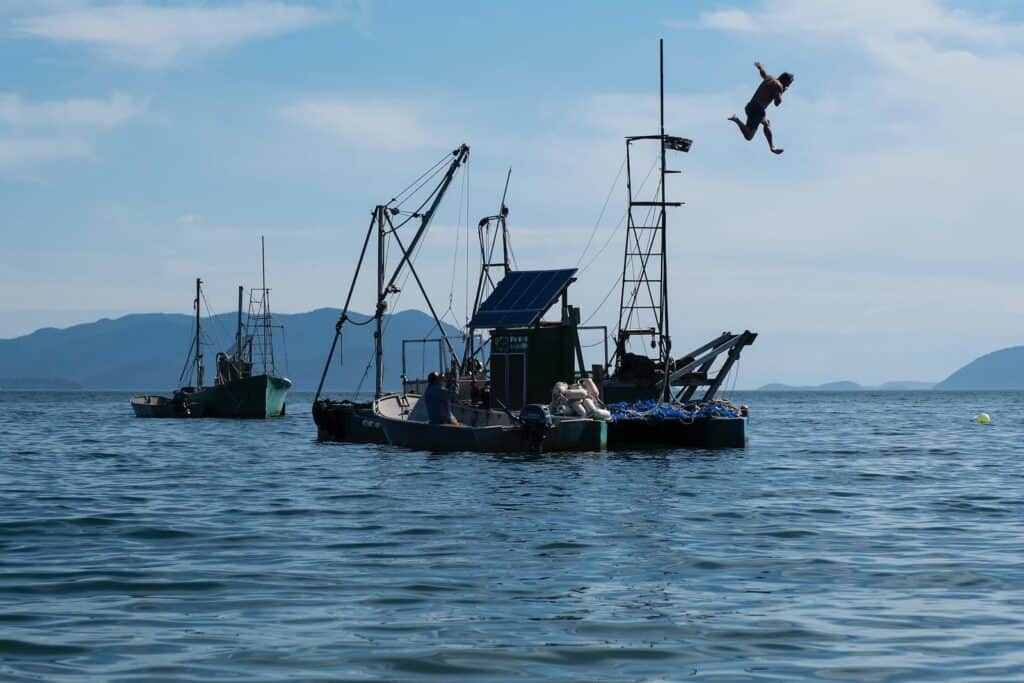 A person jumps off a fishing boat into the water, while another fishing boat floats nearby under a partly cloudy sky.