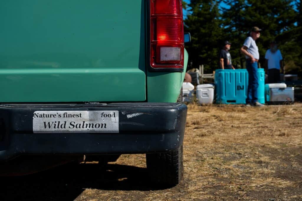 A green vehicle with a bumper sticker reading "Nature's finest food: Wild Salmon" is pictured at Legoe Bay; people and coolers are visible in the background, capturing a moment in this photo essay on reefnetters outdoors.