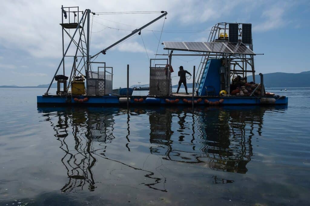A blue floating barge with equipment, solar panels, and two people stands on a calm body of water under a partly cloudy sky.